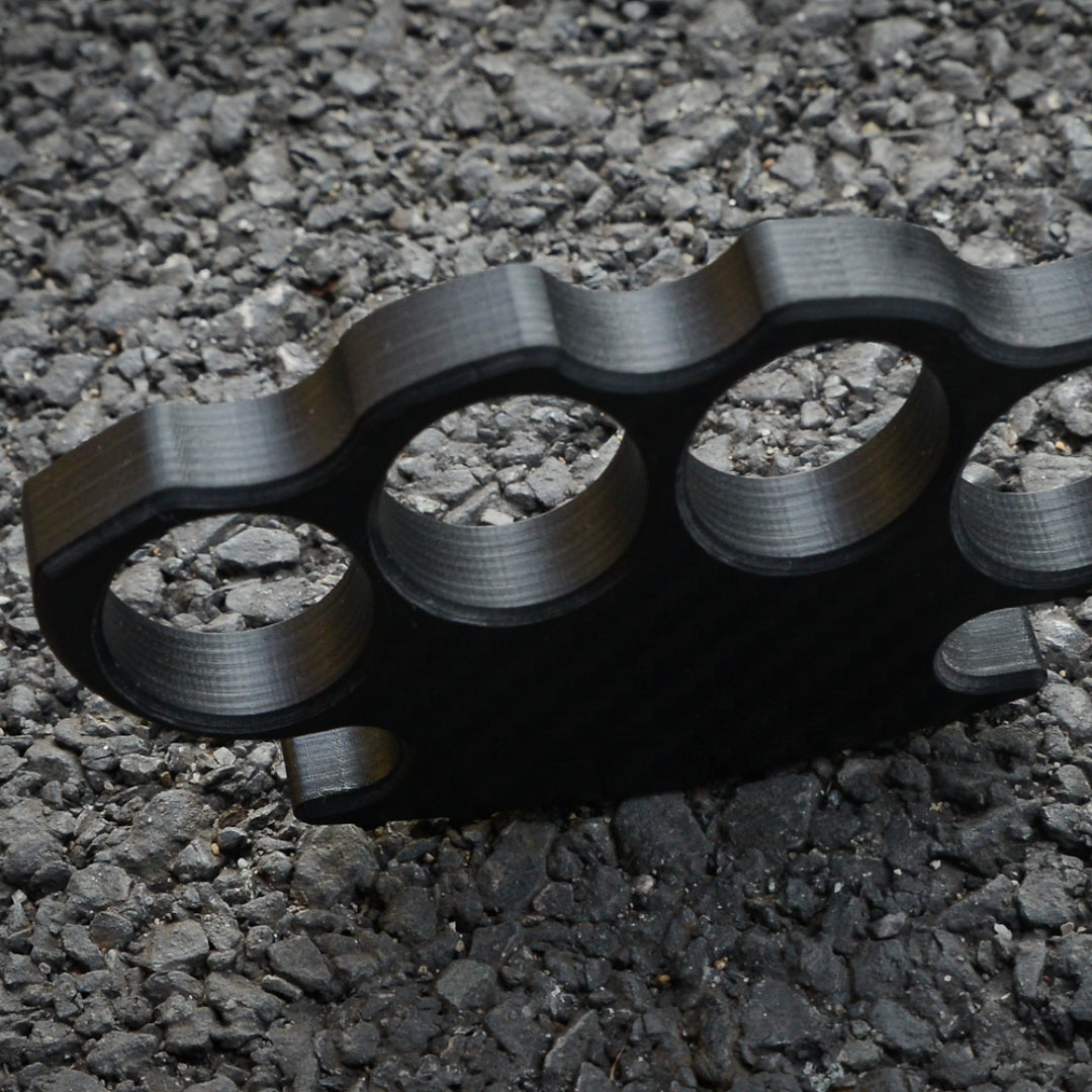 Close-up of a carbon fiber brass knuckles with four finger holes, showcasing its textured surface and structural details, placed on a gravel surface.