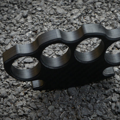 Close-up of a carbon fiber brass knuckles with four finger holes, showcasing its textured surface and structural details, placed on a gravel surface.