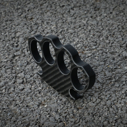 Close-up of a carbon fiber brass knuckles with four finger holes, showcasing its distinct woven pattern, placed on a gravel surface.