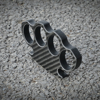 Close-up of a carbon fiber brass knuckles with four finger holes, showcasing its distinct woven pattern, placed on a gravel surface.