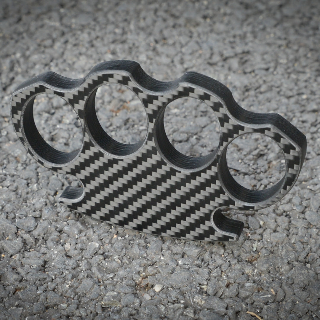 Close-up of a carbon fiber brass knuckles with four finger holes, showcasing its distinct woven carbon fiber pattern, placed on a gravel surface.
