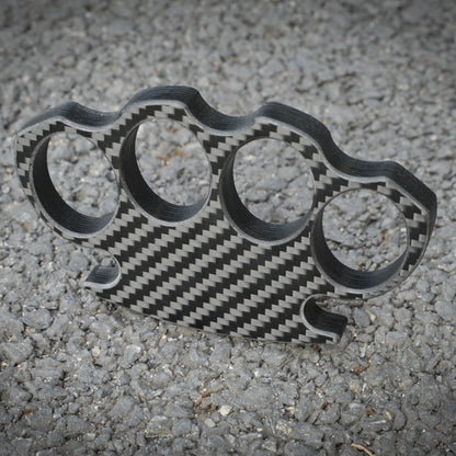 Close-up of a carbon fiber brass knuckles with four finger holes, showcasing its distinct woven carbon fiber pattern, placed on a gravel surface.
