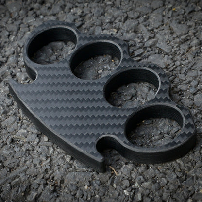 Close-up of a carbon fiber brass knuckles with four finger holes, showcasing its distinct woven carbon fiber pattern, placed on a gravel surface.