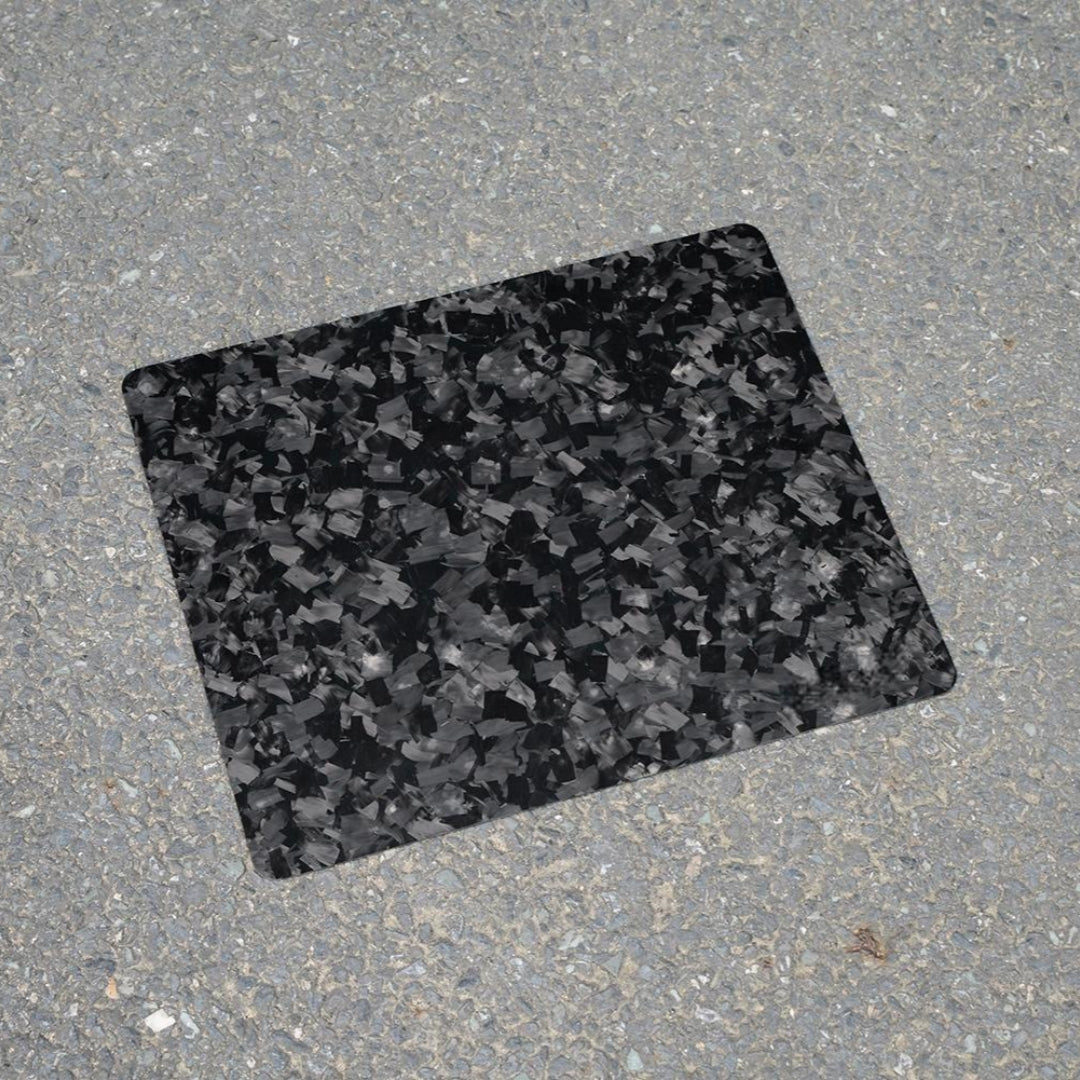 "Forged carbon fiber mouse pad featuring a textured, fragmented black and gray pattern, positioned on a rough asphalt surface."