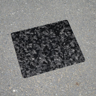 Forged carbon fiber mouse pad featuring a textured, fragmented black and gray pattern, positioned on a rough asphalt surface