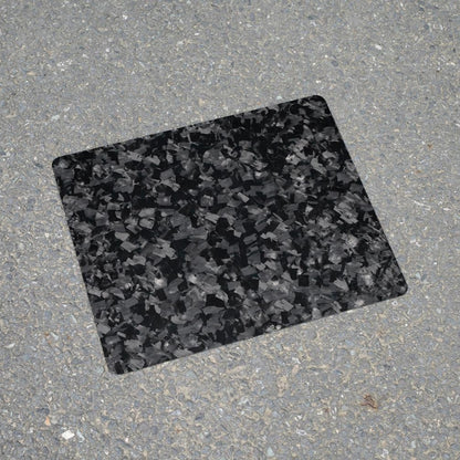 "Forged carbon fiber mouse pad featuring a textured, fragmented black and gray pattern, positioned on a rough asphalt surface."
