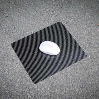A white wireless mouse resting on a 3K carbon fiber mouse pad with a textured black pattern, placed on a rough asphalt surface.