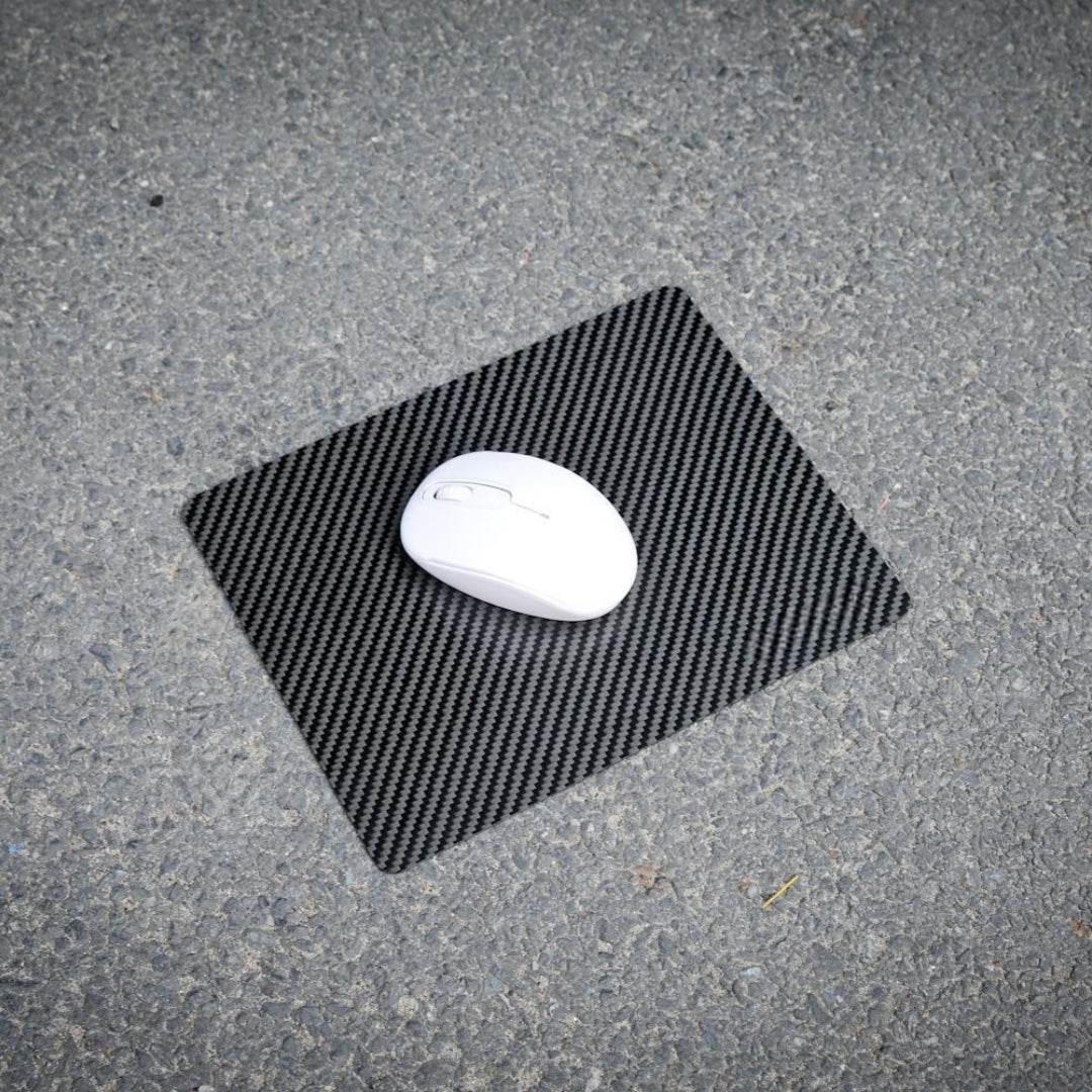 A white wireless mouse resting on a 3K carbon fiber mouse pad with a textured black pattern, placed on a rough asphalt surface.