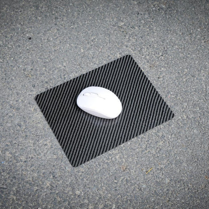 A white wireless mouse resting on a 3K carbon fiber mouse pad with a textured black pattern, placed on a rough asphalt surface.