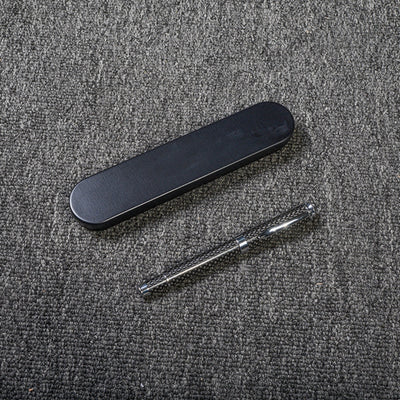 Close-up of a carbon fiber ballpoint pen with a distinct woven texture, accompanied by a sleek black metal case, displayed on a gray textured fabric surface.