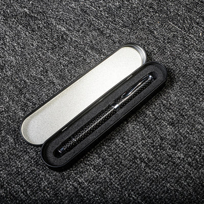 Close-up of a carbon fiber ballpoint pen with a distinct woven texture, nestled in a black metal case with foam lining and a silver interior