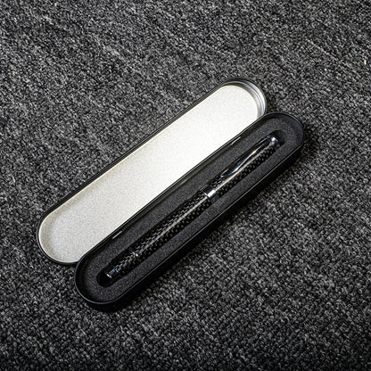 Close-up of a carbon fiber ballpoint pen with a distinct woven texture, nestled in a black metal case with foam lining and a silver interior, displayed on a gray textured fabric surface.
