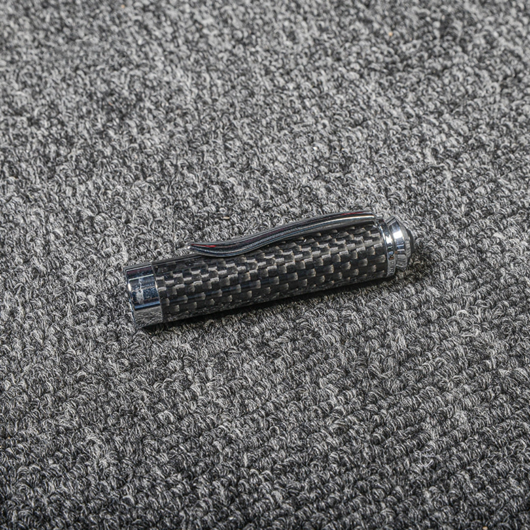 Close-up of a carbon fiber ballpoint pen cap with a distinct woven texture, metallic accents, and a clip, displayed on a gray textured fabric surface.