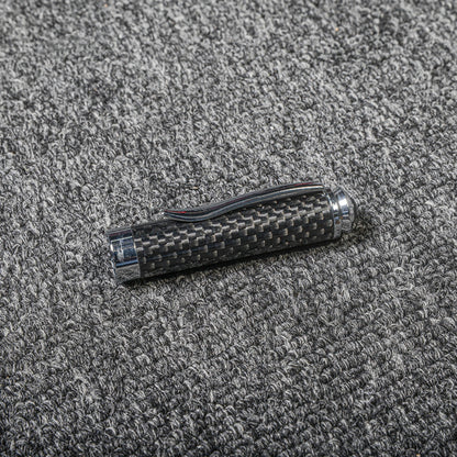 Close-up of a carbon fiber ballpoint pen cap with a distinct woven texture, metallic accents, and a clip, displayed on a gray textured fabric surface.