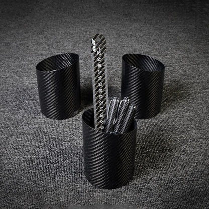 Three carbon fiber pen holders with a distinct woven pattern, where the central one contains a carbon fiber ruler and several pens, displayed on a gray textured surface.