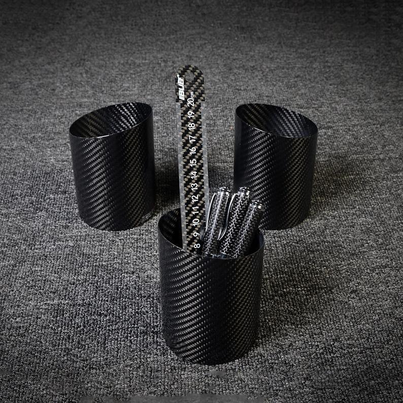 Three carbon fiber pen holders with a distinct woven pattern, where the central one contains a carbon fiber ruler and several pens, displayed on a gray textured surface.