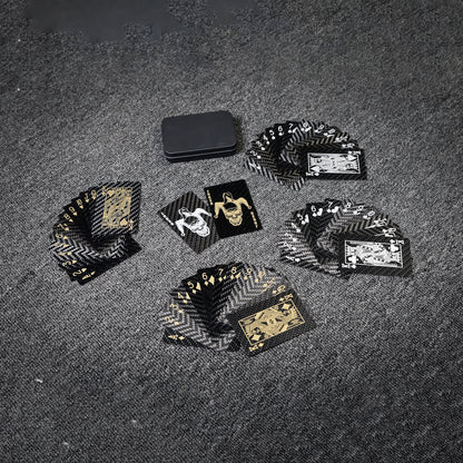 Close-up of carbon fiber playing cards with black and gold/white detailing, along with a black tin case, arranged on a gray textured surface.