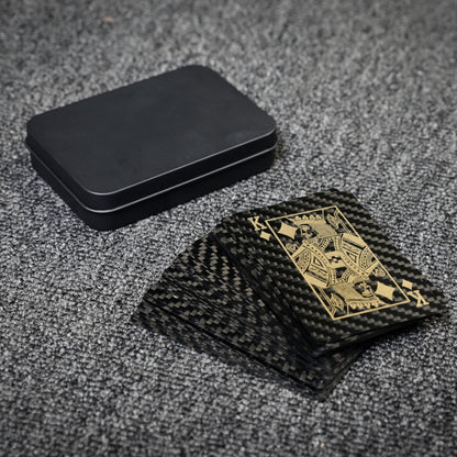 Close-up of carbon fiber playing cards with gold detailing (featuring the King of Diamonds) and a black tin case, laid on a gray textured surface.