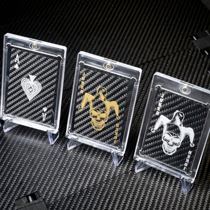 Close-up of three carbon fiber playing cards (Ace of Spades, gold Joker, and white Joker) in clear protective cases, displayed on a carbon fiber textured surface.