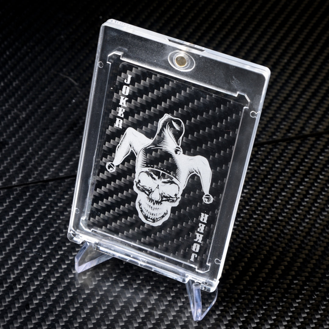 Close-up of a carbon fiber Joker playing card with a white skull jester design, encased in a clear protective case, displayed on a carbon fiber textured surface.
