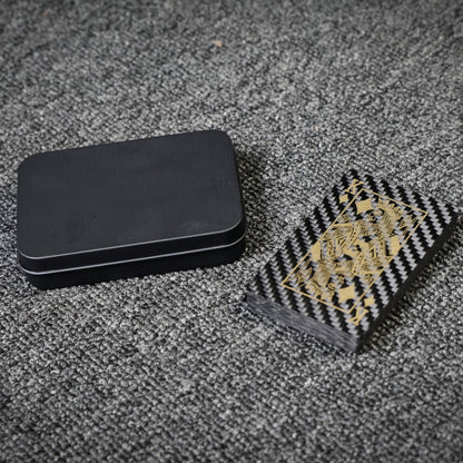 Close-up of a deck of carbon fiber playing cards with gold detailing (featuring the King card) and a black tin case, laid on a gray textured surface.