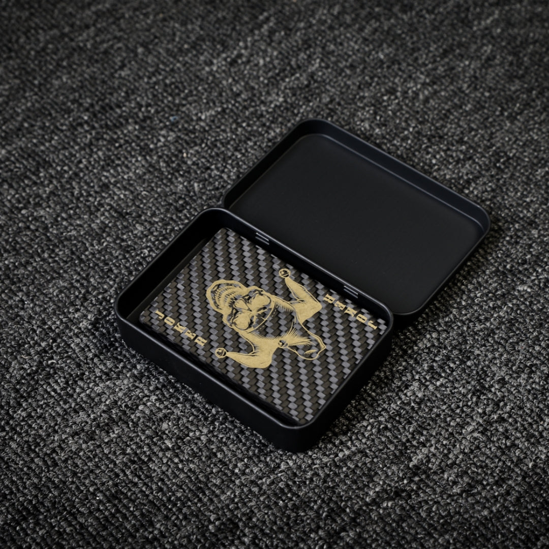 Close-up of a deck of carbon fiber playing cards with a gold Joker design and carbon fiber texture, placed in an open black tin case on a gray textured surface.