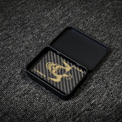 Close-up of a deck of carbon fiber playing cards with a gold Joker design and carbon fiber texture, placed in an open black tin case on a gray textured surface.