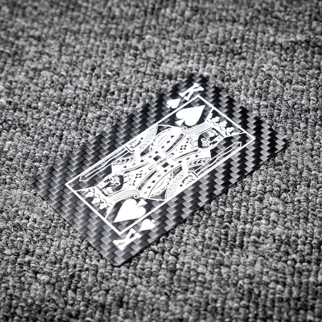 Close-up of a carbon fiber playing card featuring the King of Spades with white detailing, showcasing the distinct carbon fiber texture, laid on a gray textured surface.