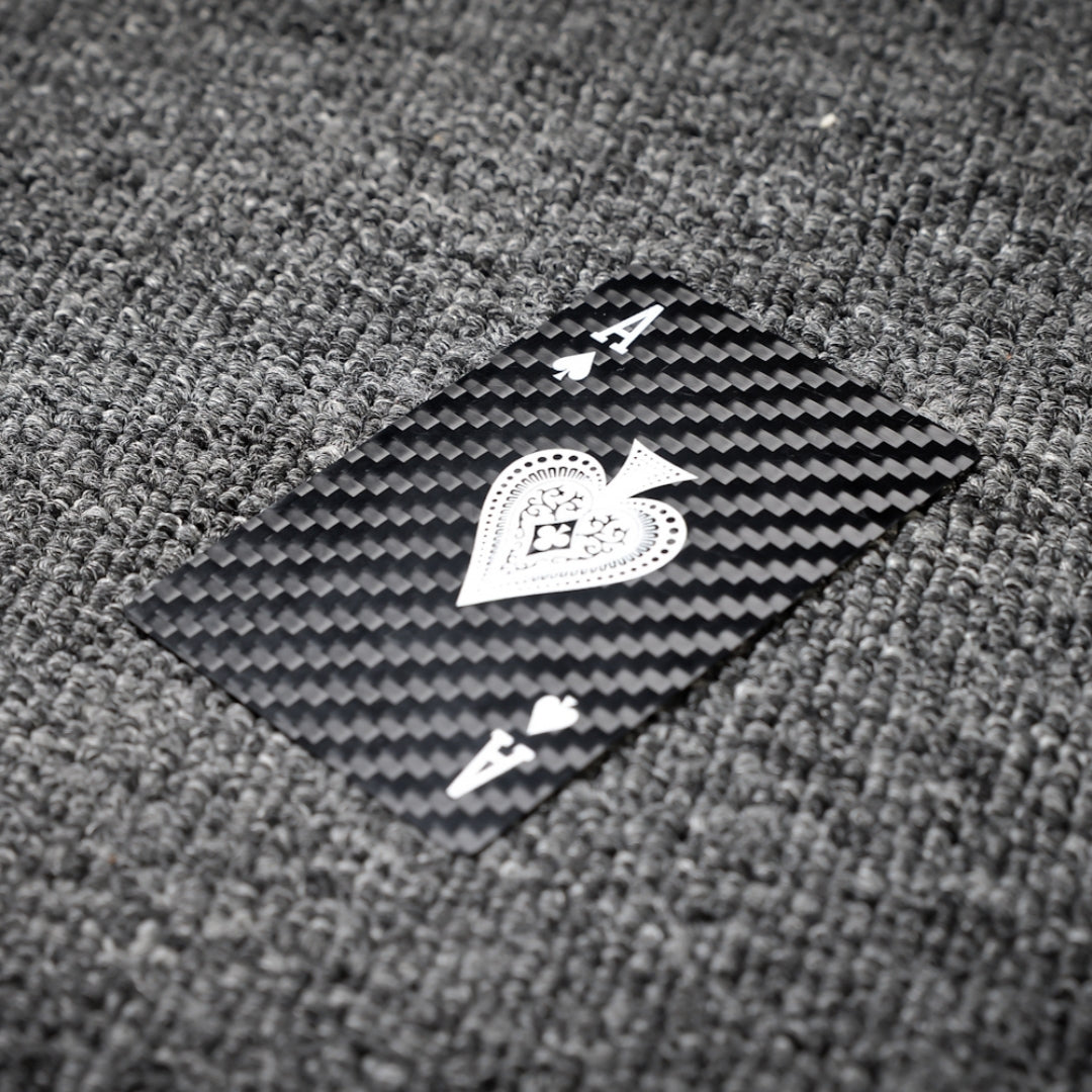 Close-up of a carbon fiber playing card (Ace of Spades) with white detailing, showcasing the distinct carbon fiber textured pattern, laid on a gray textured surface.
