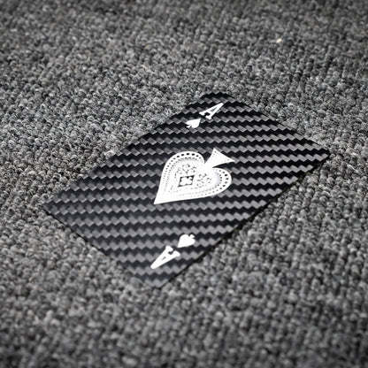 Close-up of a carbon fiber playing card (Ace of Spades) with white detailing, showcasing the distinct carbon fiber textured pattern, laid on a gray textured surface.
