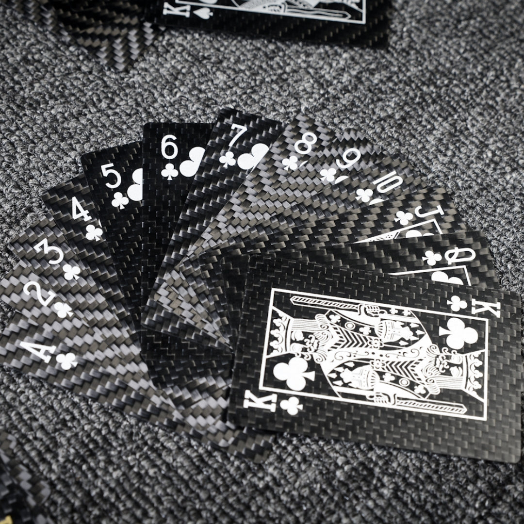 Close-up of carbon fiber playing cards fanned out with Club suit cards (Ace to King), showcasing crisp white detailing and the distinct carbon fiber weave pattern, placed on a gray textured surface.