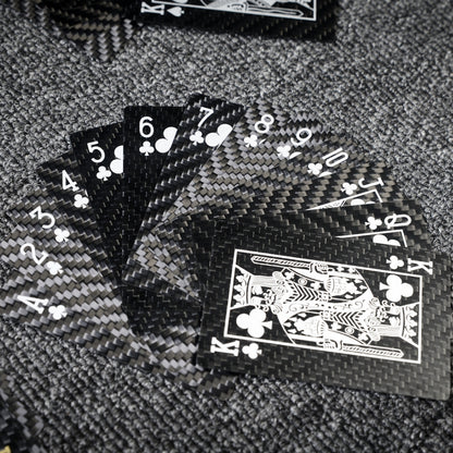 Close-up of carbon fiber playing cards fanned out with Club suit cards (Ace to King), showcasing crisp white detailing and the distinct carbon fiber weave pattern, placed on a gray textured surface.