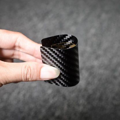 Close-up of a carbon fiber playing card with gold detailing, bent to showcase its flexibility, held by a hand against a gray background, highlighting the distinct carbon fiber weave pattern.