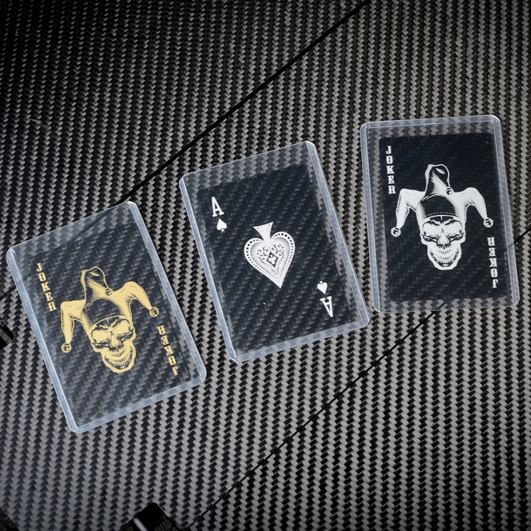 Close-up of three carbon fiber playing cards (two Jokers with gold and white skull-jester designs, and an Ace of Spades) in clear protective cases, displayed on a carbon fiber textured surface.