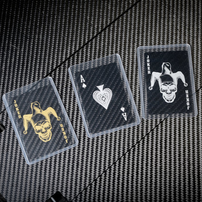 Close-up of three carbon fiber playing cards (two Jokers with gold and white skull-jester designs, and an Ace of Spades) in clear protective cases, displayed on a carbon fiber textured surface.