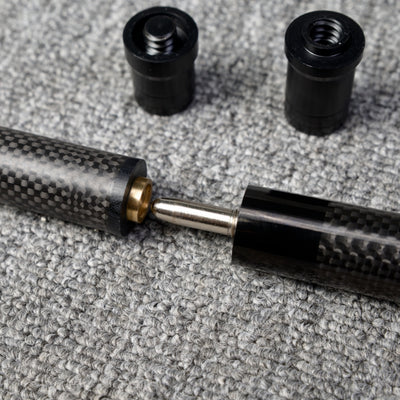 Close-up detail of a two-piece carbon fiber pool cue with a textured carbon fiber pattern, along with two black accessories