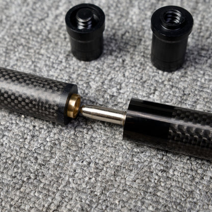 Close-up detail of a two-piece carbon fiber pool cue with a textured carbon fiber pattern, showcasing the metal joint and brass fitting connection, along with two black accessories, placed on a gray textured surface.