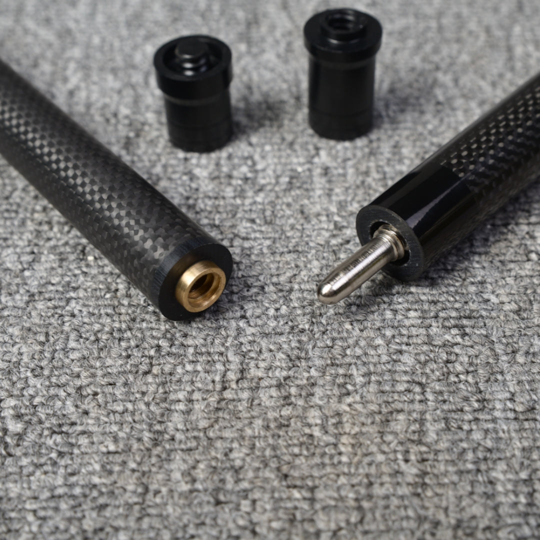 Close-up detail of a two-piece carbon fiber pool cue with a textured carbon fiber pattern, showcasing the metal joint, brass fitting, and two black accessories, placed on a gray textured surface.