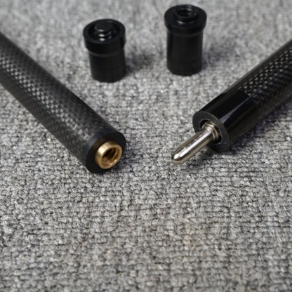 Close-up detail of a two-piece carbon fiber pool cue with a textured carbon fiber pattern, showcasing the metal joint, brass fitting, and two black accessories, placed on a gray textured surface.
