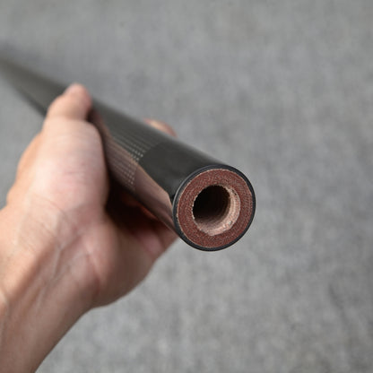 Close-up of a hand holding a carbon fiber pool cue, showcasing the cross-sectional detail with a textured carbon fiber exterior and its inner structure, against a gray background.