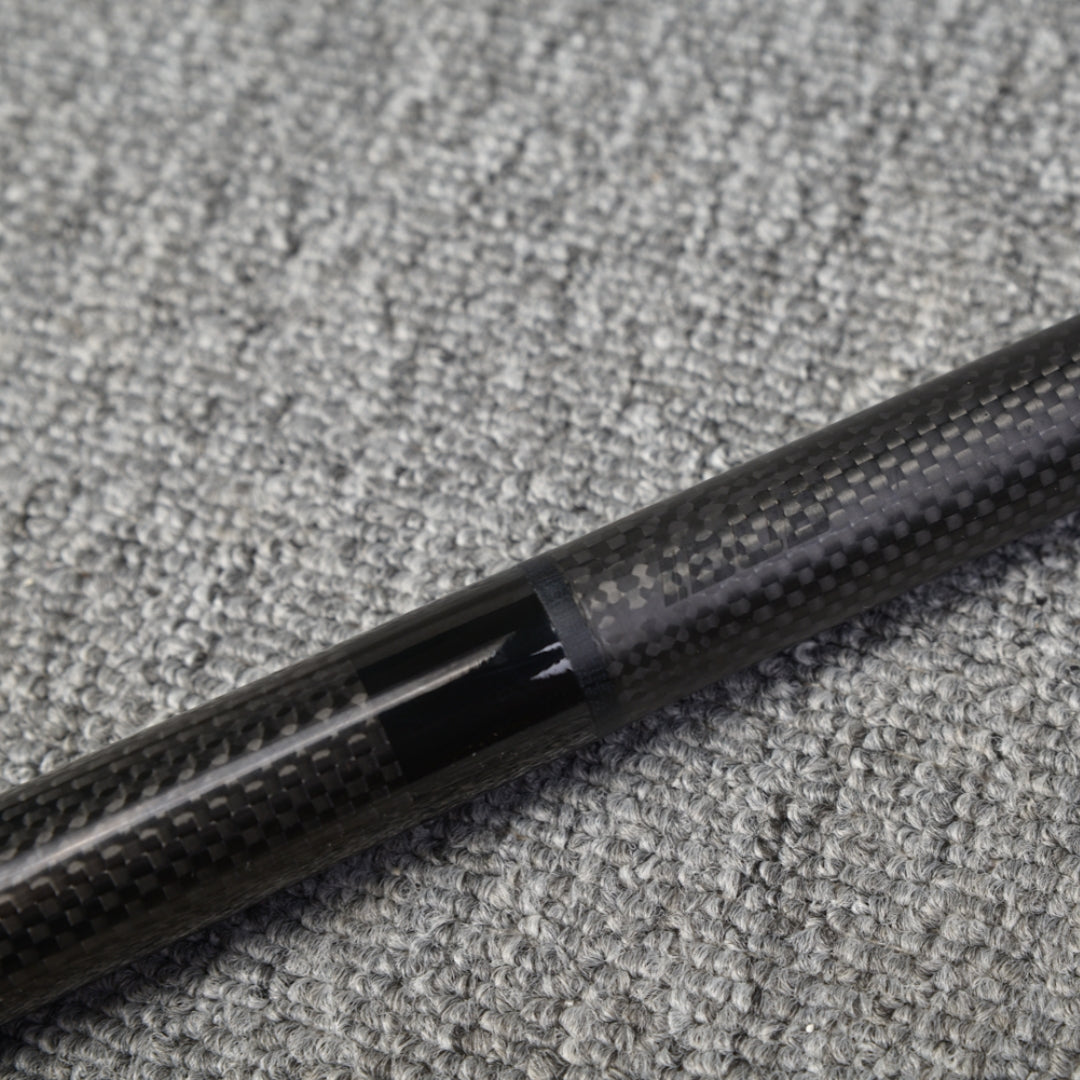 Close-up detail of a two-piece carbon fiber pool cue showcasing the connection area with a textured carbon fiber pattern, placed on a gray textured surface.