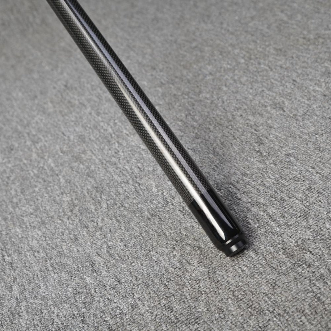 Close-up detail of the end of a carbon fiber pool cue, showcasing a smooth black finish at the tip and a textured carbon fiber pattern on the shaft, resting on a gray textured surface.