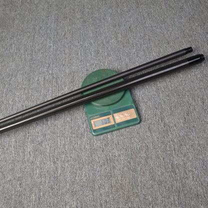 Close-up of two carbon fiber pool cues placed on a digital scale displaying a weight reading, set against a gray textured surface.