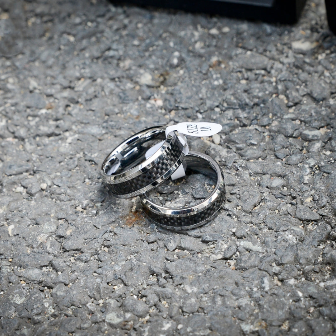 Two rings featuring 3K carbon fiber detailing and shiny edges, with one ring attached to a "SIZE 10" tag, resting on a rough gravel surface.