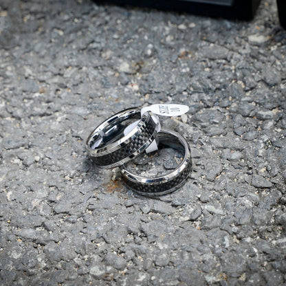 Two rings featuring 3K carbon fiber detailing and shiny edges, with one ring attached to a "SIZE 10" tag, resting on a rough gravel surface.