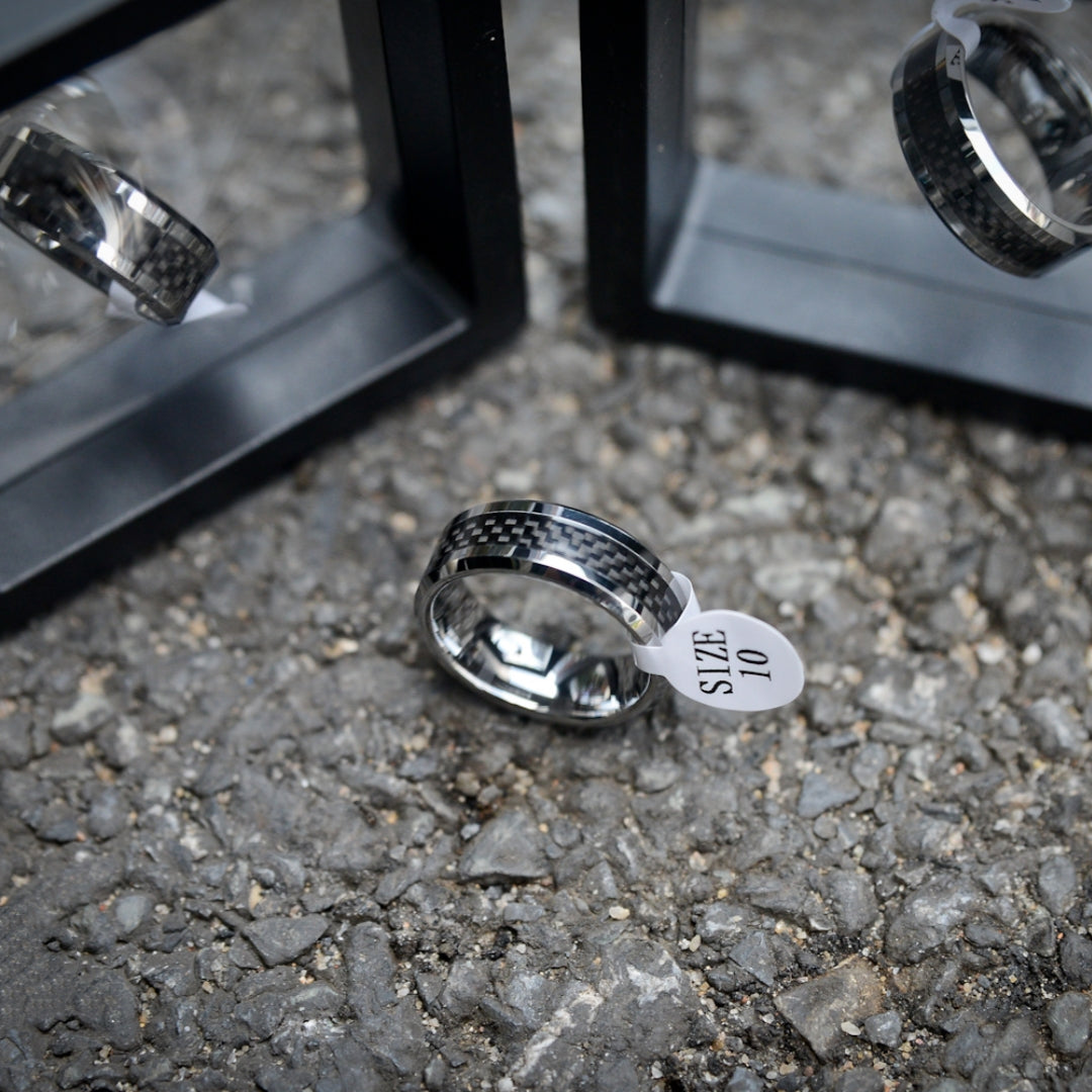 A 3K carbon fiber ring with a shiny metallic band, attached to a white size tag marked "SIZE 10", placed on a rough gravel surface, with black display stands and reflections of the ring visible in the background.