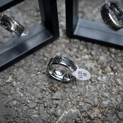 A 3K carbon fiber ring with a shiny metallic band, attached to a white size tag marked "SIZE 10", placed on a rough gravel surface, with black display stands and reflections of the ring visible in the background.