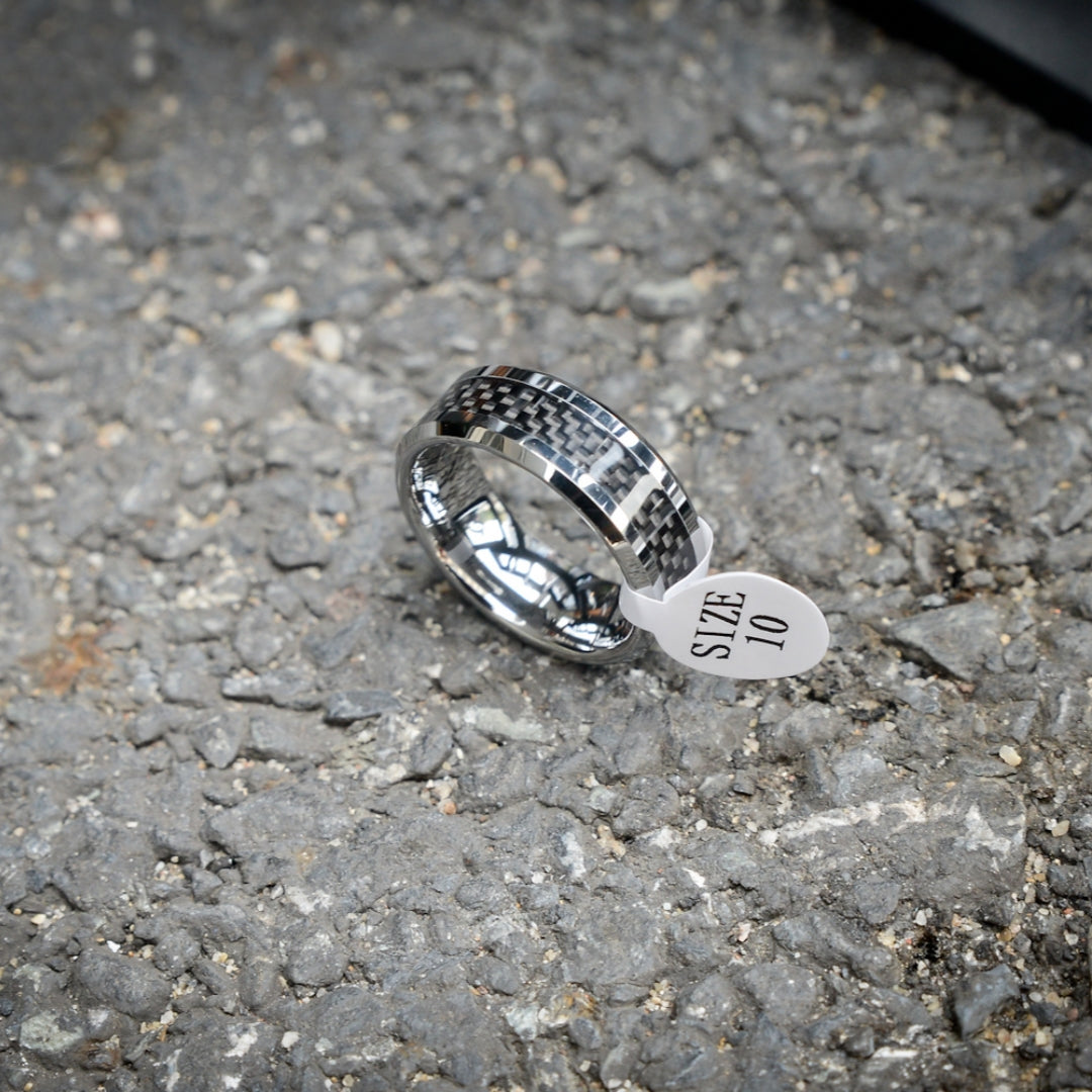 A 3K carbon fiber ring with a shiny metallic band, attached to a white size tag that reads "SIZE 10", resting on a rough gravel surface.