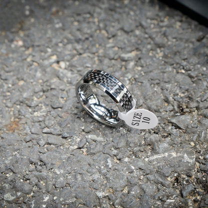 A 3K carbon fiber ring with a shiny metallic band, attached to a white size tag that reads "SIZE 10", resting on a rough gravel surface.