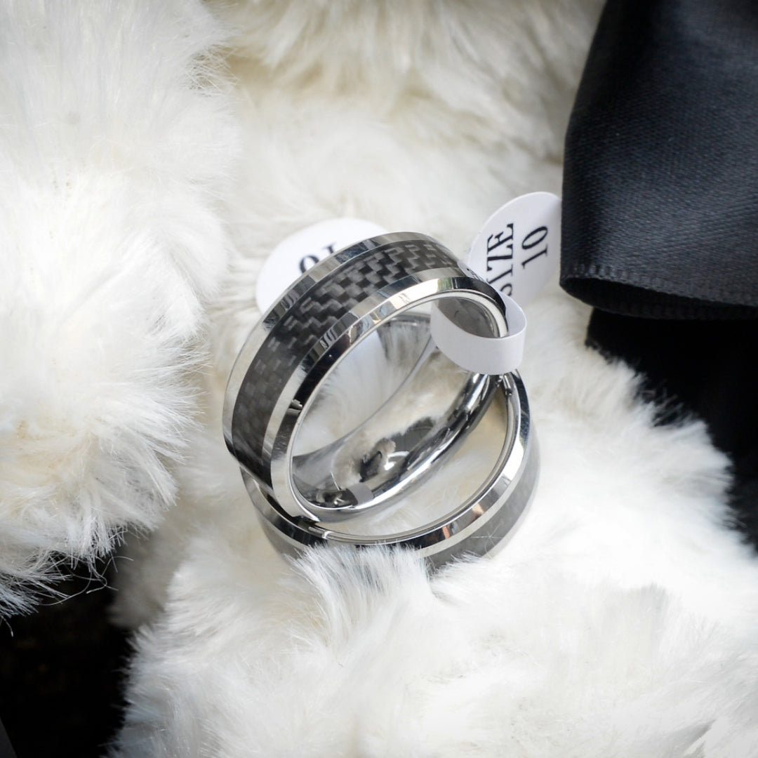 Two rings with 3K carbon fiber detailing and shiny metallic bands, each attached to a white size tag marked "SIZE 10", resting on a soft, fluffy white surface with black fabric visible nearby.